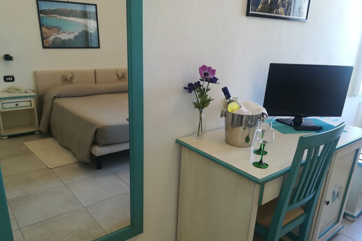 A room with a bed, a desk with a computer, a vase with flowers, and a bottle.