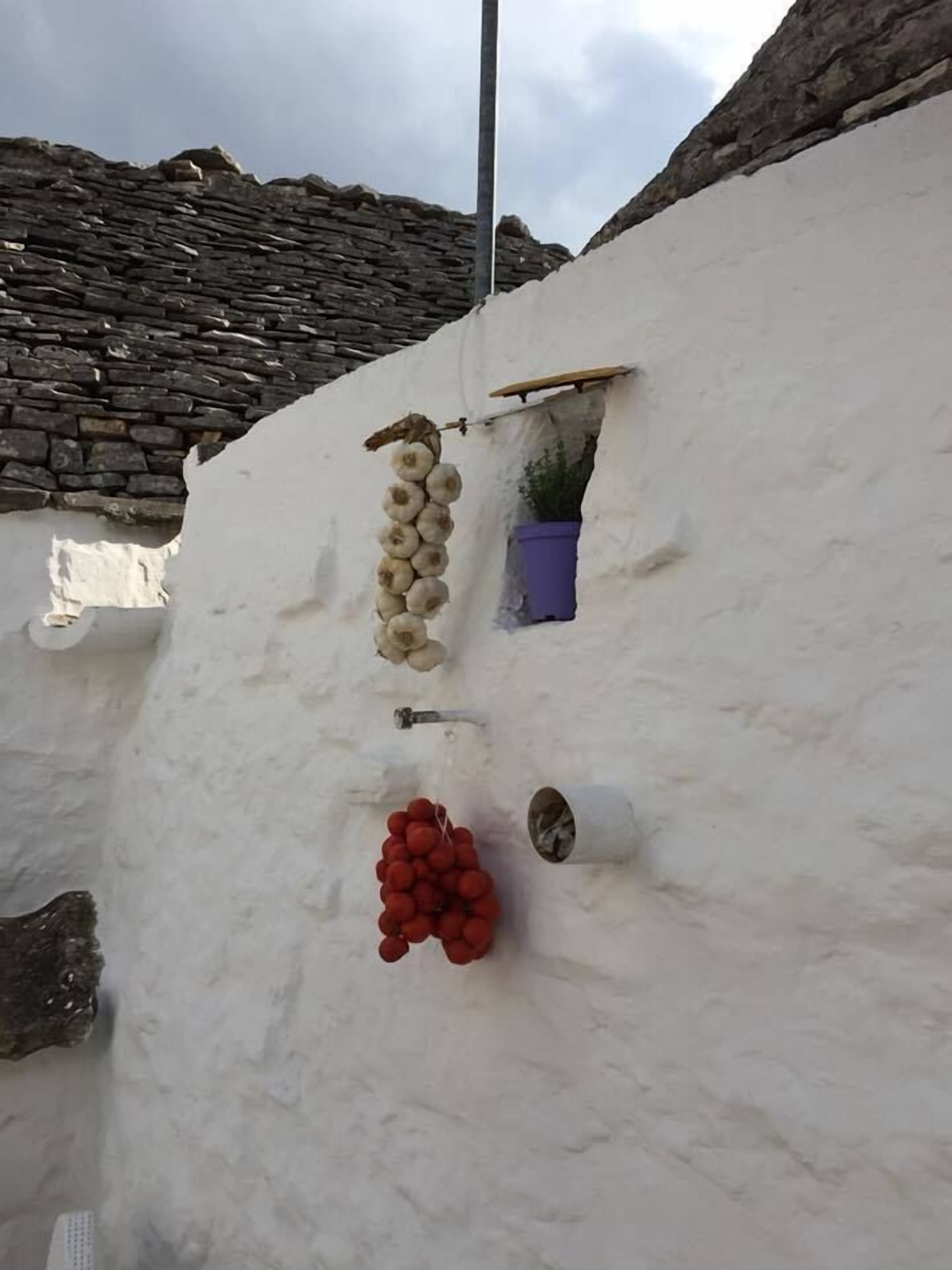 A stone wall with a white wall adjacent to it, a small window with a plant, and a hanging bunch of red grapes.