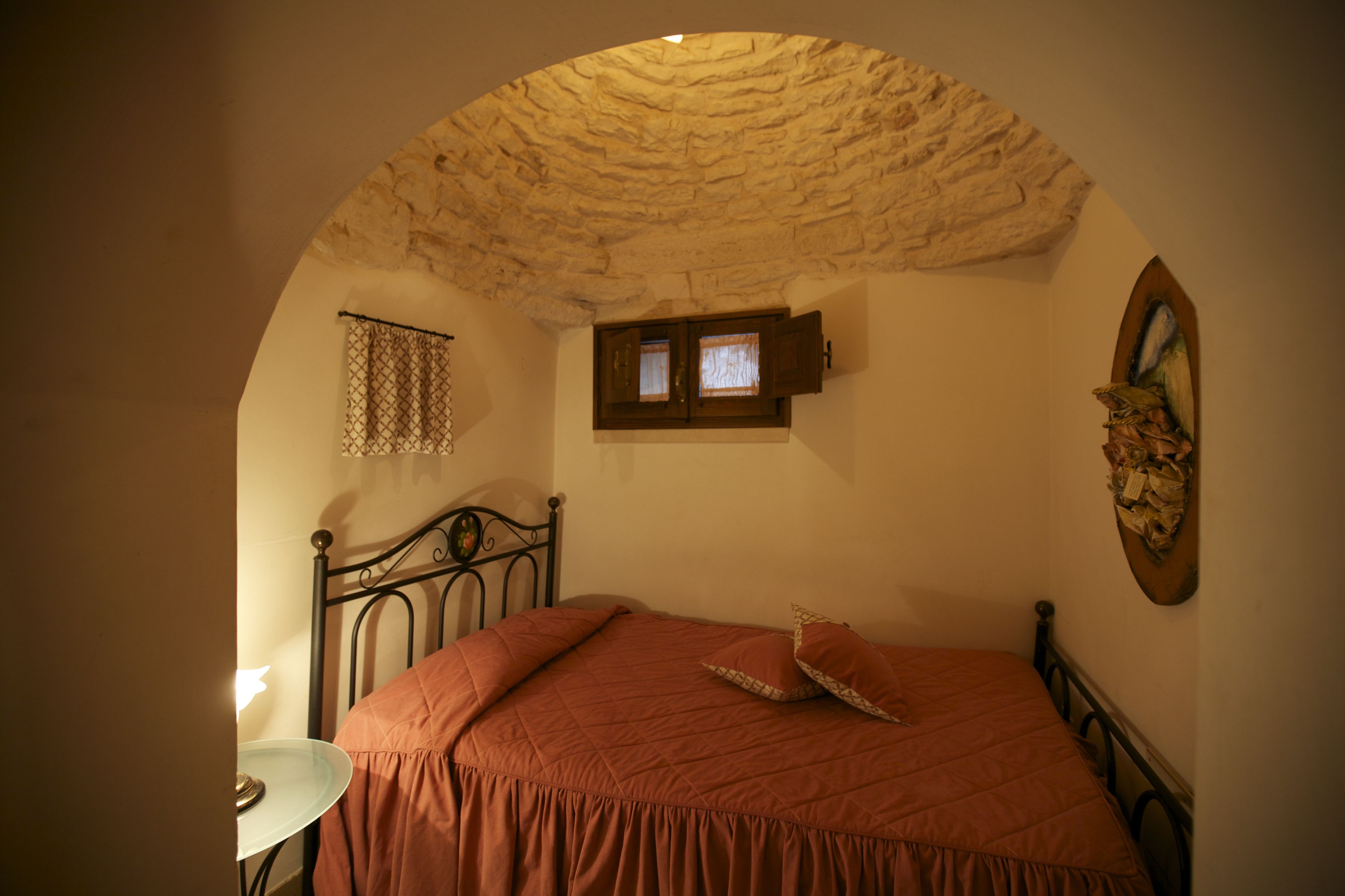 Ein Schlafzimmer mit Steindecke, einem Bett mit roter Bettdecke, einem kleinen Fenster mit Rollläden und einem an der Wand hängenden Gemälde.