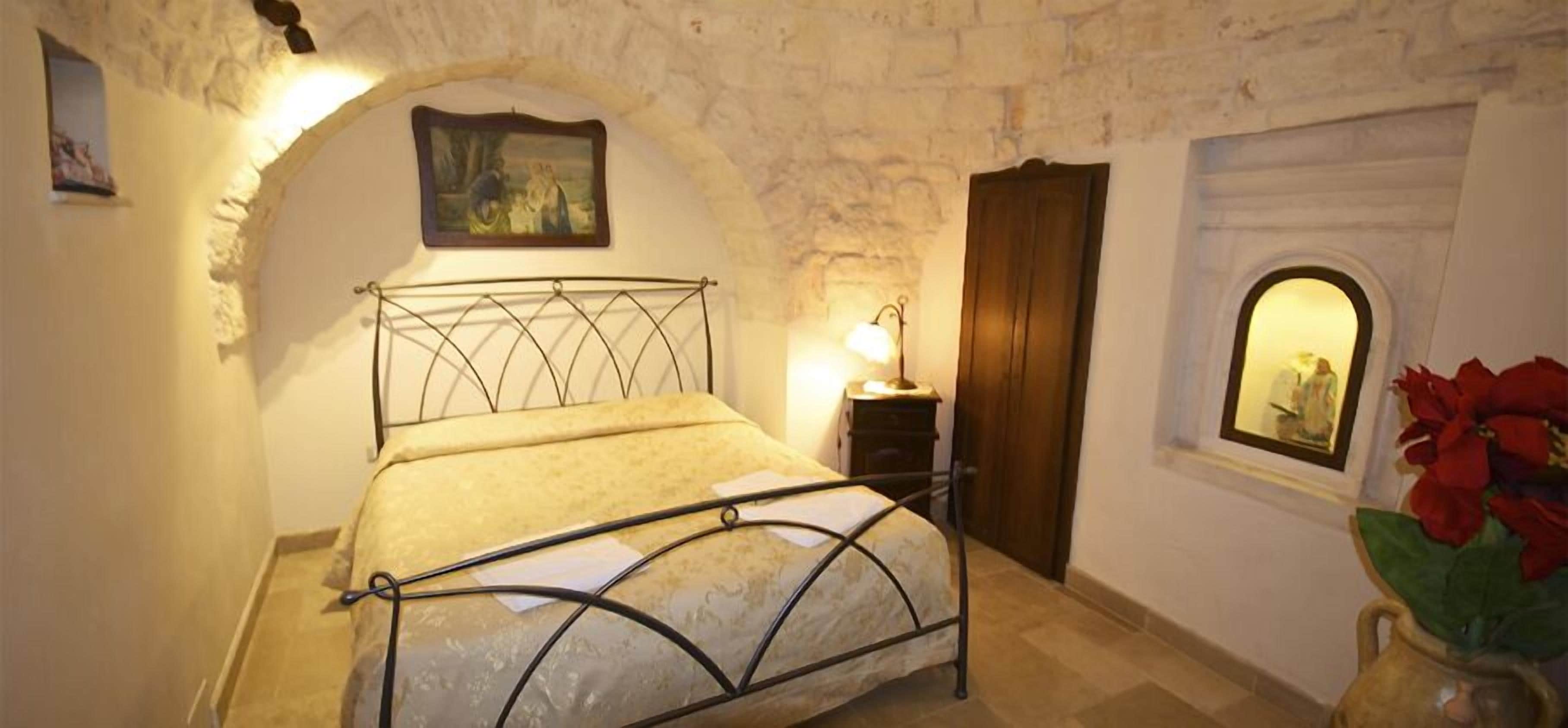 Une chambre avec un mur en pierre, un lit avec un cadre en métal, une table de chevet avec une lampe et une fenêtre donnant sur un beau paysage.
