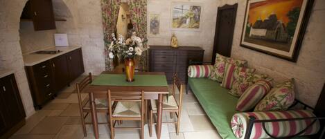 A cozy room with a dining table, chairs, a sofa with floral cushions, a green bed, and a painting on the wall.