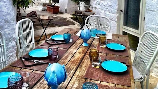A dining area with a long wooden table set for four, surrounded by white chairs. There are potted plants and a small table with a chair outside the main area. - Trulli e Puglia (Alberobello)