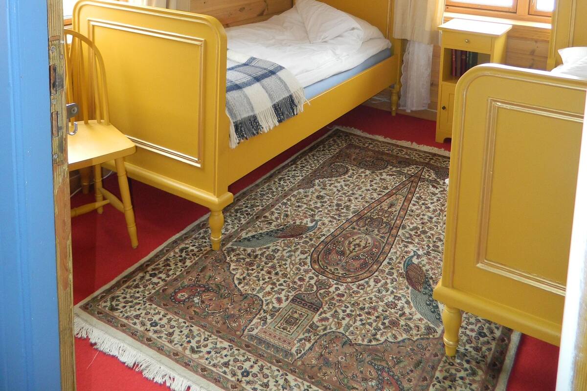 Una habitació amb un mur amb panells de fusta, un llit amb capçalera groga, una cadira de fusta, una taula petita i un penjador.