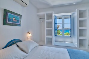 A bedroom with a bed, a wall-mounted light, a window with a view of the sea, and a framed picture on the wall. - Blue Dolphin Antiparos Villas and Suites (Antiparos)