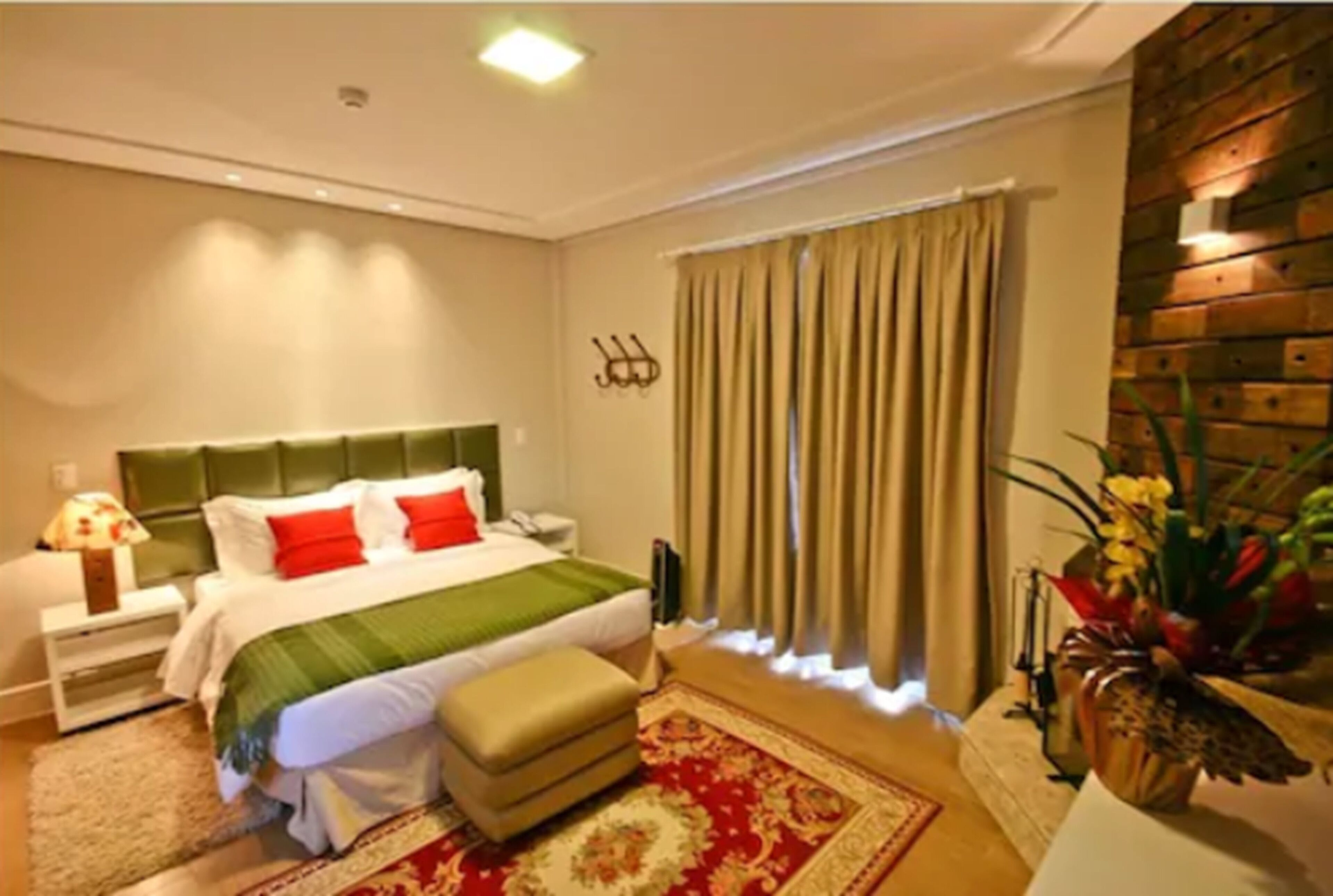 A hotel room with a bed, a nightstand, a lamp, a window with curtains, a wall-mounted shelf with decorative items, and a wooden accent wall.