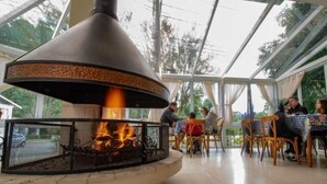 Ein Restaurant mit einem zentralen Kamin, Sitzgelegenheiten im Freien und Blick auf Bäume.