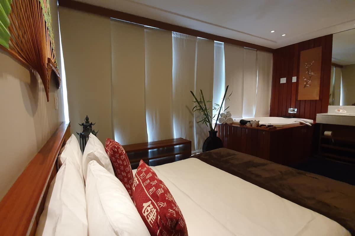 Une chambre d’hôtel avec un lit, un banc en bois, une petite table avec des serviettes, une plante et une salle de bain équipée d’un lavabo et d’un miroir.