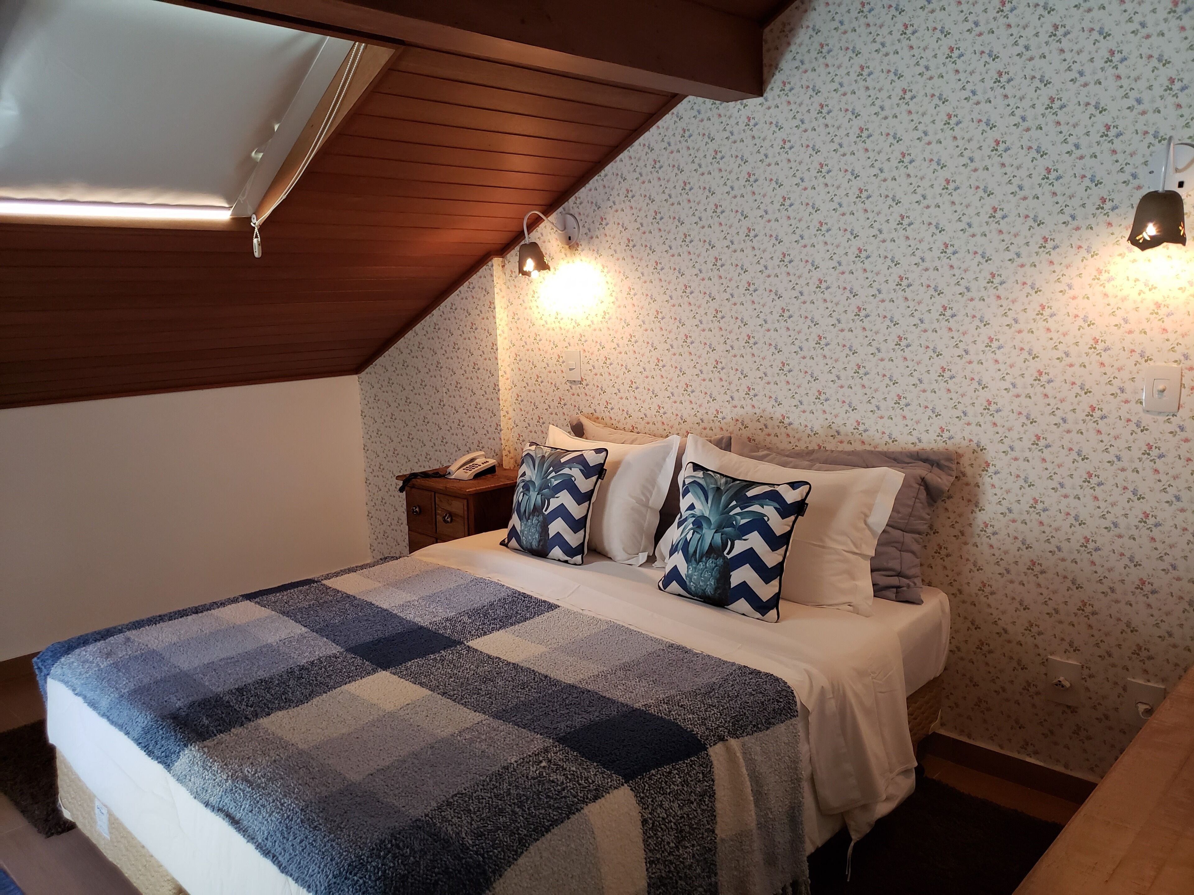 A bedroom with a checkered bedspread, two pillows with a pineapple design, a wooden nightstand with a phone, and a wall-mounted light fixture.