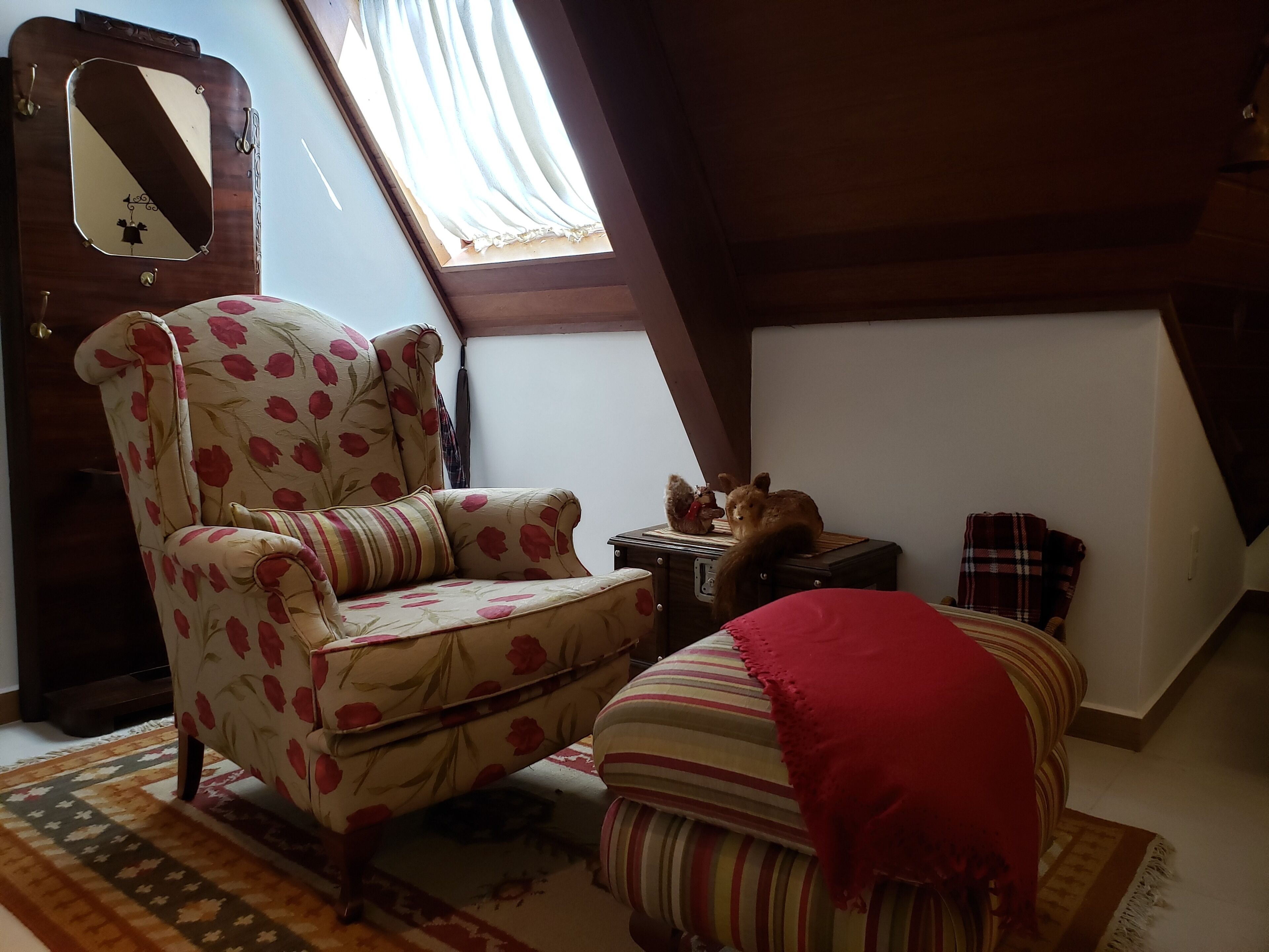 A cozy attic room with a floral armchair, a small ottoman, a vintage radio, and a cat.