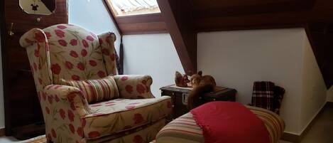 A cozy attic room with a floral armchair, a small ottoman, a vintage radio, and a cat.