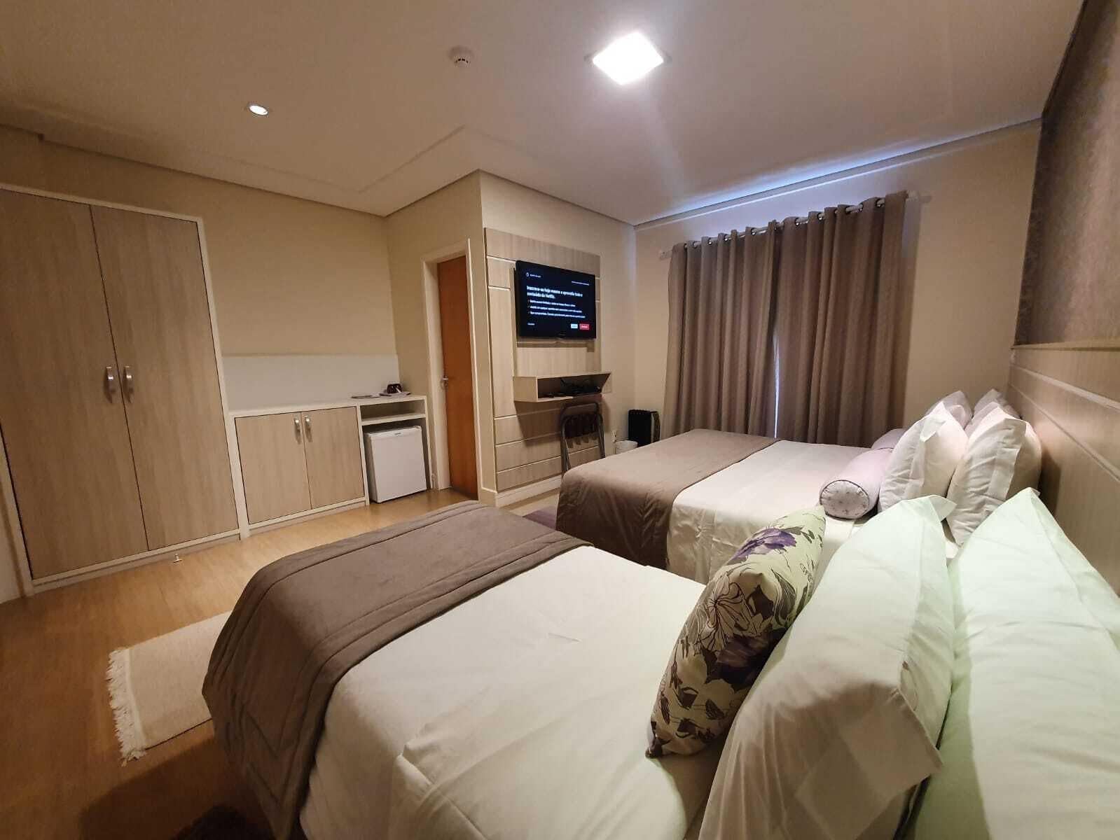Ein Hotelzimmer mit zwei Betten, einer kleinen Küchenzeile, einem Flachbildfernseher und einem großen Fenster mit Vorhängen.
