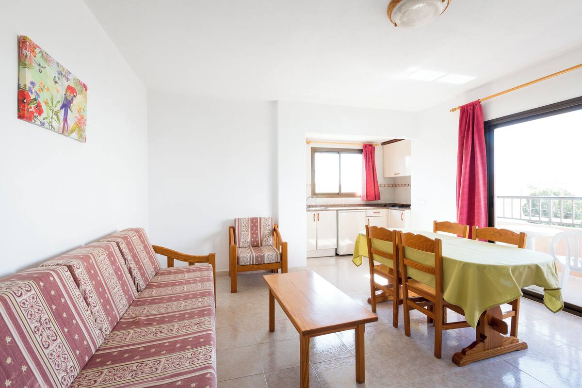 Ein Wohnzimmer mit einer Couch, einem Holztisch und einem Esstisch mit Stühlen. Im Hintergrund ist eine Küche mit einem Fenster und einem Balkon zu sehen.