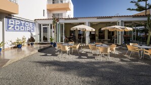 An outdoor cafe area with tables and chairs, a building with a sign reading "APTOS ZODIAC", and potted plants. - Zodiac Apartamentos (Sant Josep de sa Talaia)