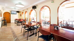 A restaurant with multiple tables and chairs, a television mounted on the wall, and arched windows allowing natural light. - Zodiac Apartamentos (Sant Josep de sa Talaia)