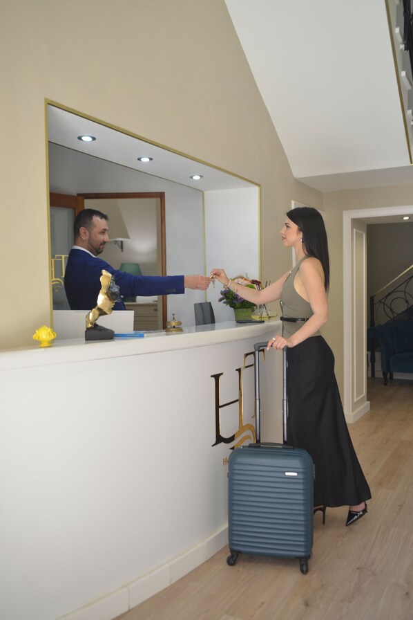 Un mostrador de recepción con un espejo, una maleta azul y una mujer entregando llaves a un hombre con traje azul.