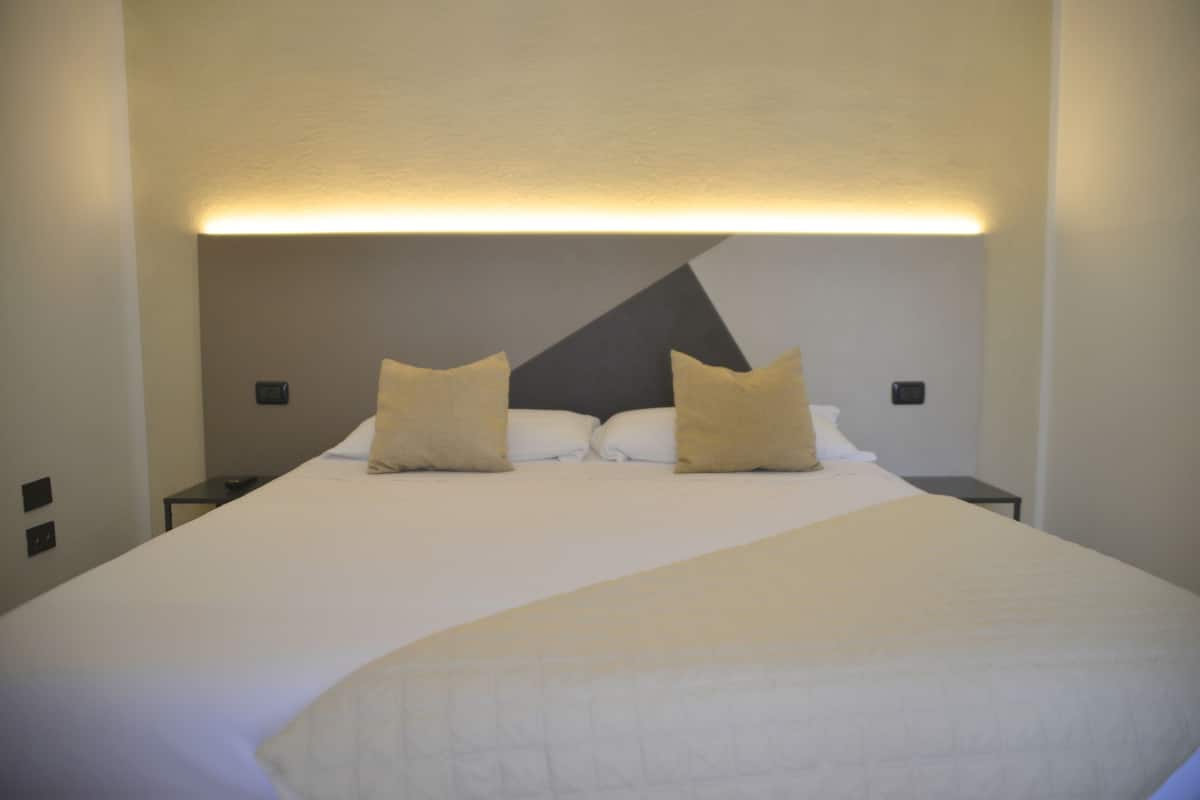 Un lit bien fait, avec du linge de lit blanc et deux oreillers beige, une table de chevet avec une lampe, et un luminaire fixé au mur au-dessus du lit.
