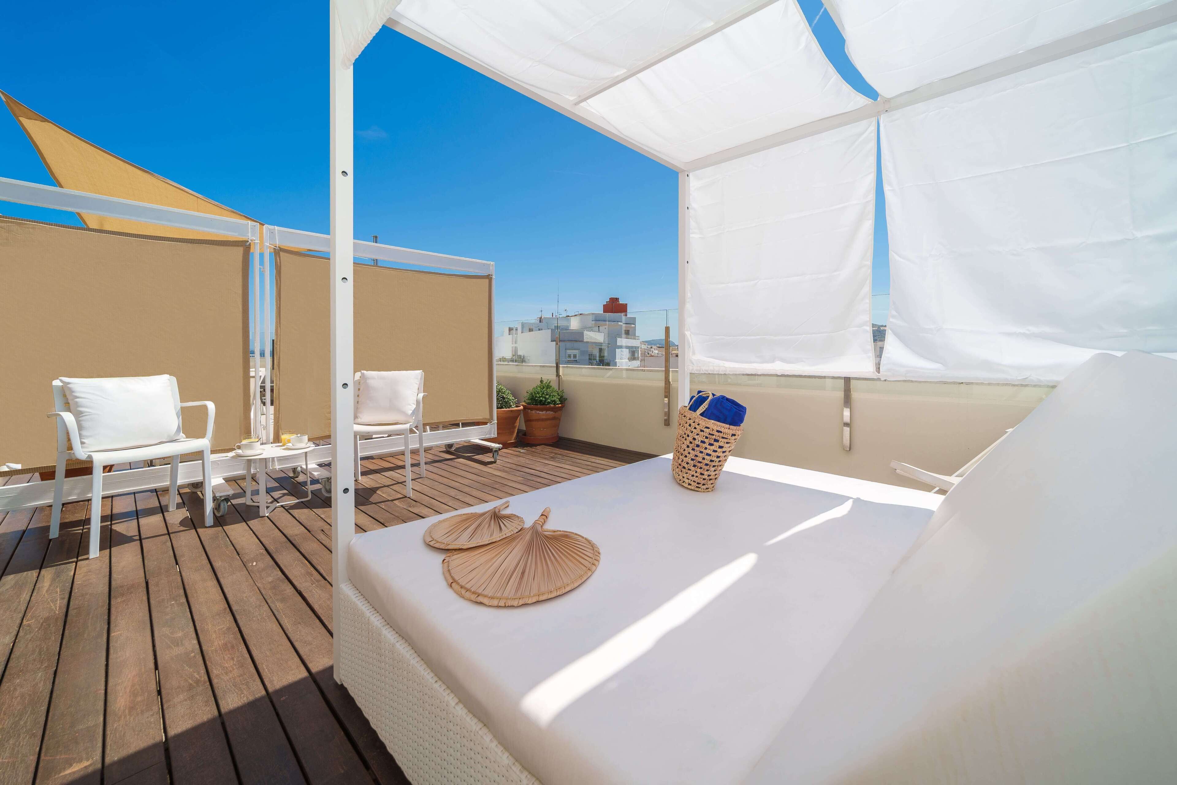 A sunlit balcony with a white bed, two white chairs, a small table, and a potted plant.