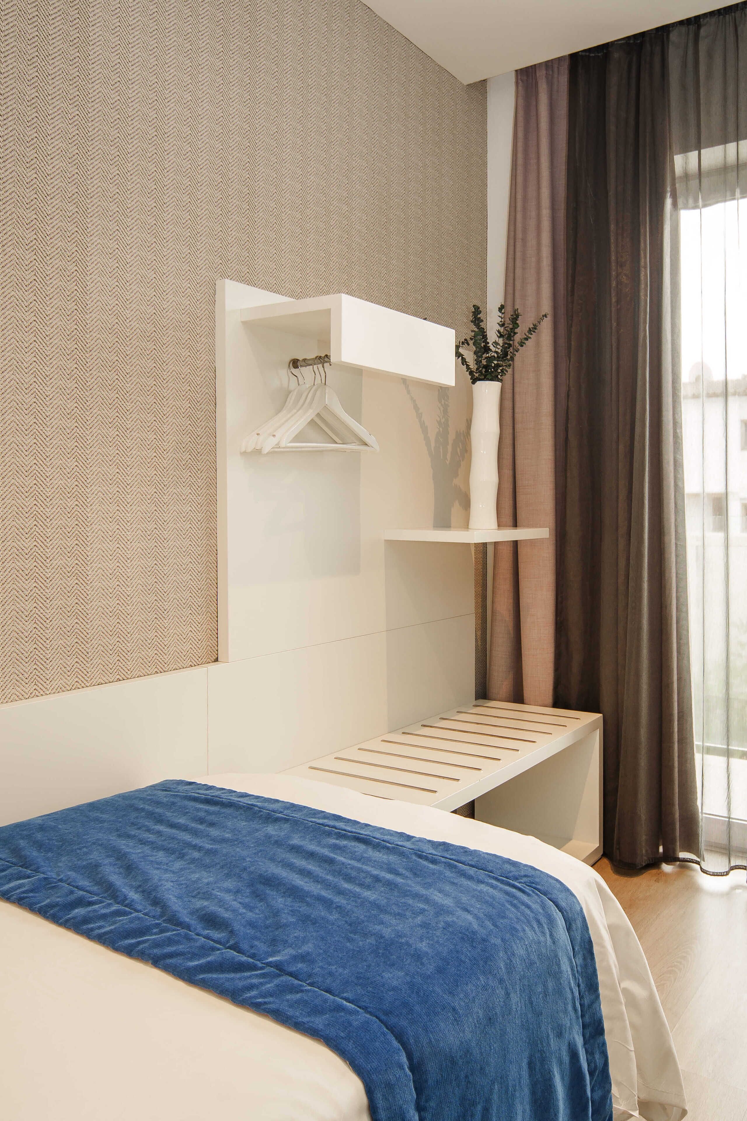 A modern hotel room with a bed, a white wall-mounted shelf with a hanging garment, and a window with curtains.