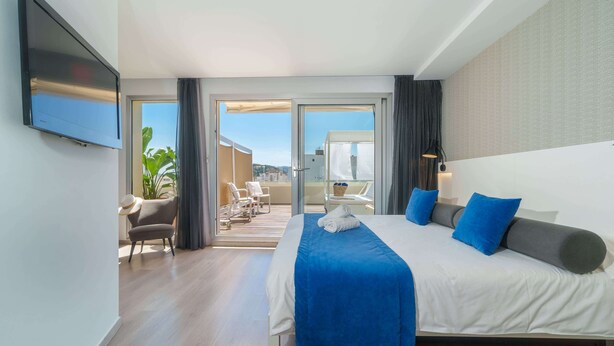 A modern hotel room with a large bed, a flat-screen TV, a balcony with seating, and a view of the city. - Mar Calma Hotel (Pollenca)