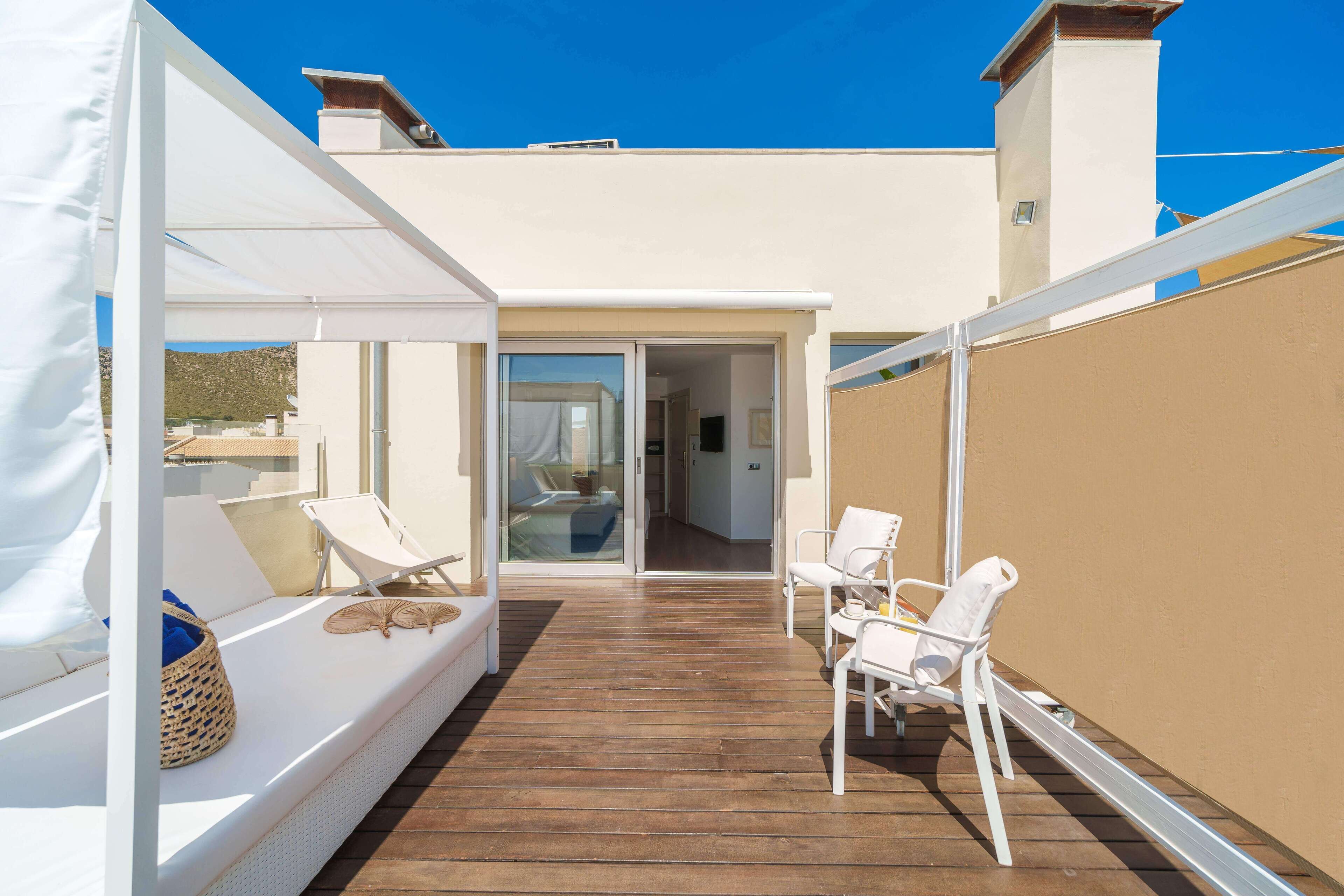 A sunlit outdoor seating area with a wooden deck, white lounge chairs, and a covered seating area with cushions.