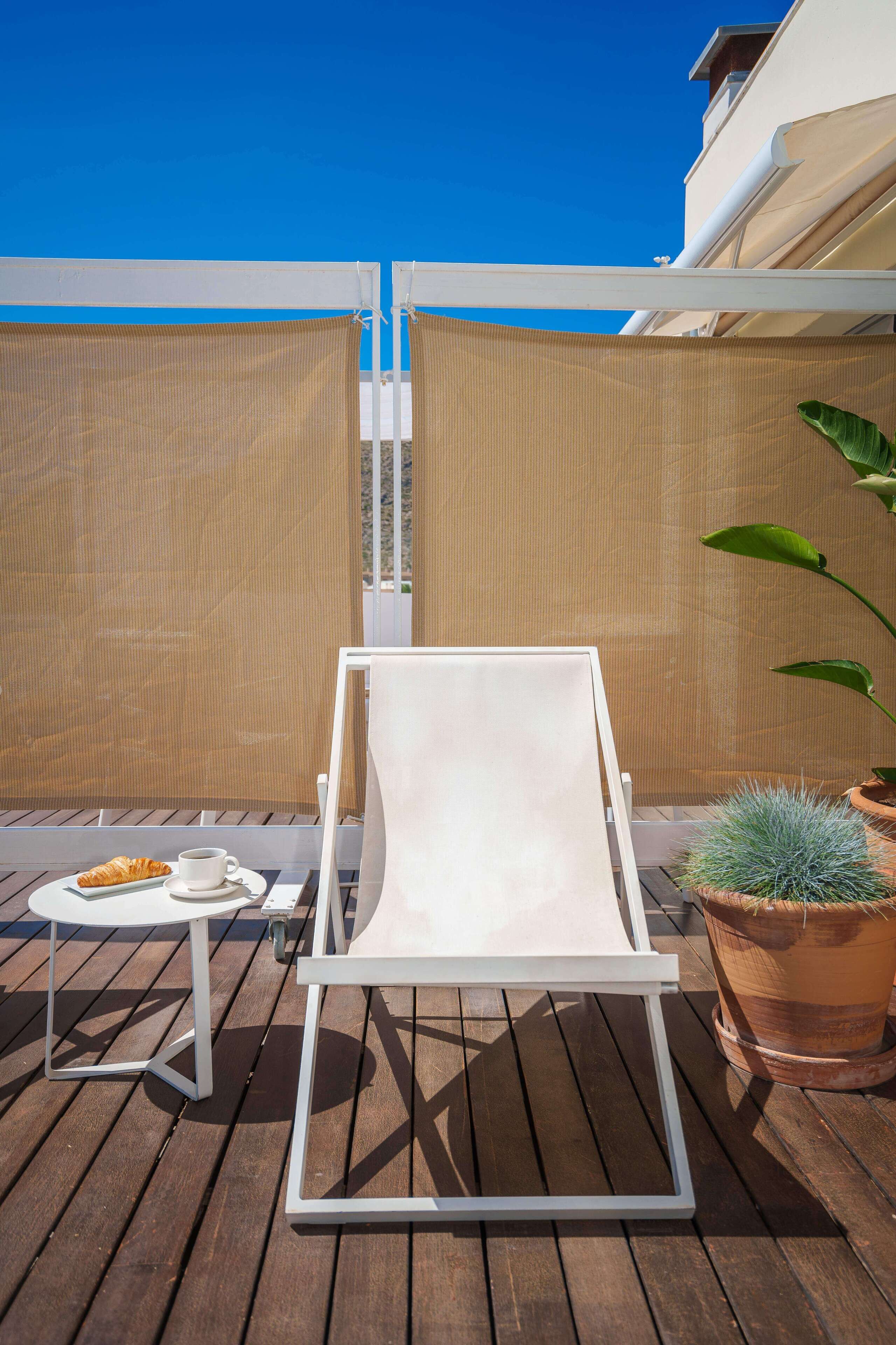 A sunlit outdoor seating area with a white lounge chair, a small table with a croissant and a cup, and a potted plant.