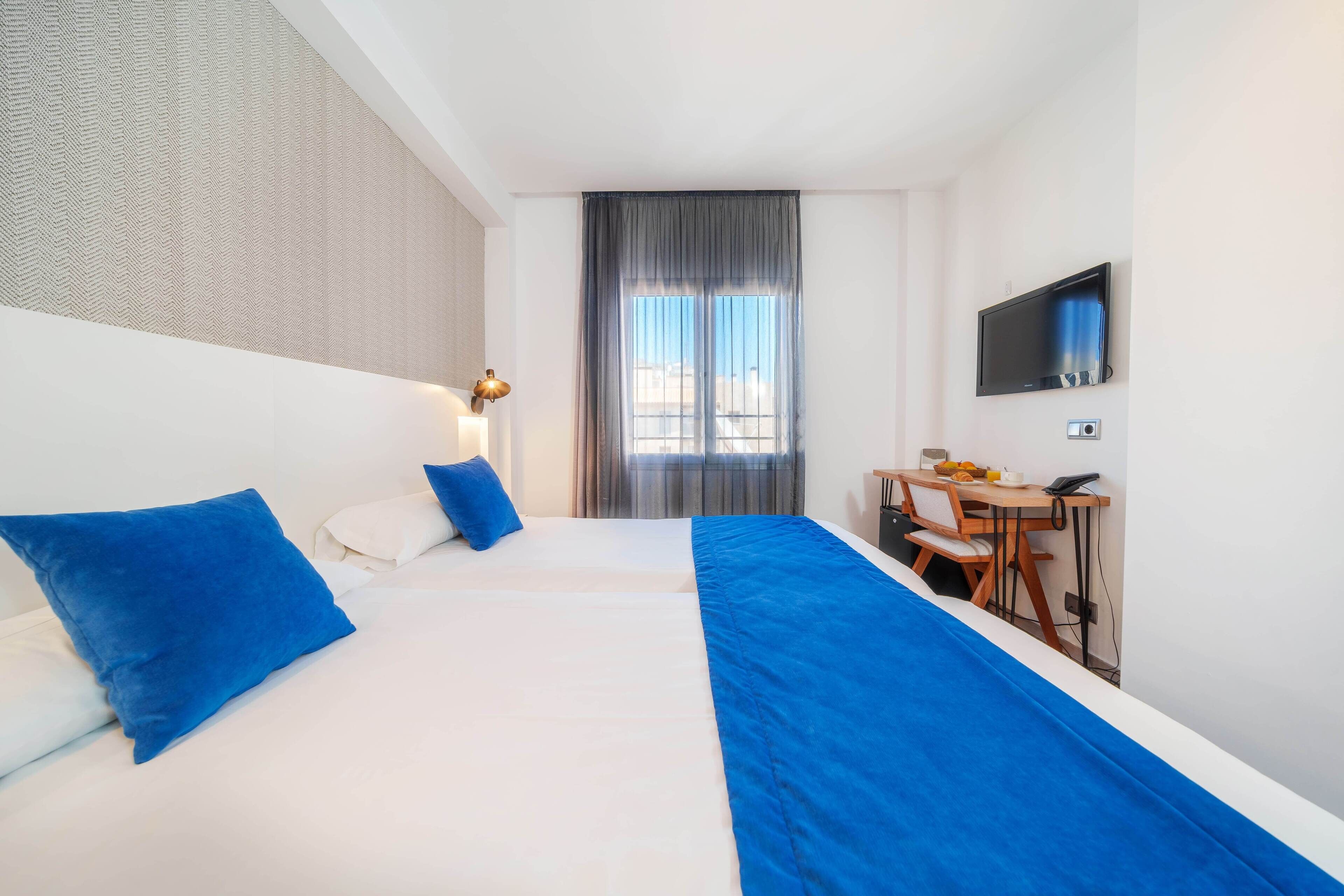 Ein Hotelzimmer mit einem großen Bett, zwei blauen Kissen, einem Schreibtisch mit Stuhl, einem Flachbildfernseher und einem Fenster mit Vorhängen.