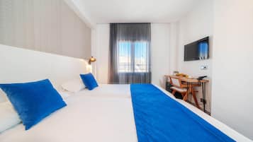 Habitación de hotel con una cama grande, dos almohadas azules, un escritorio con una silla, un televisor de pantalla plana y una ventana con cortinas.