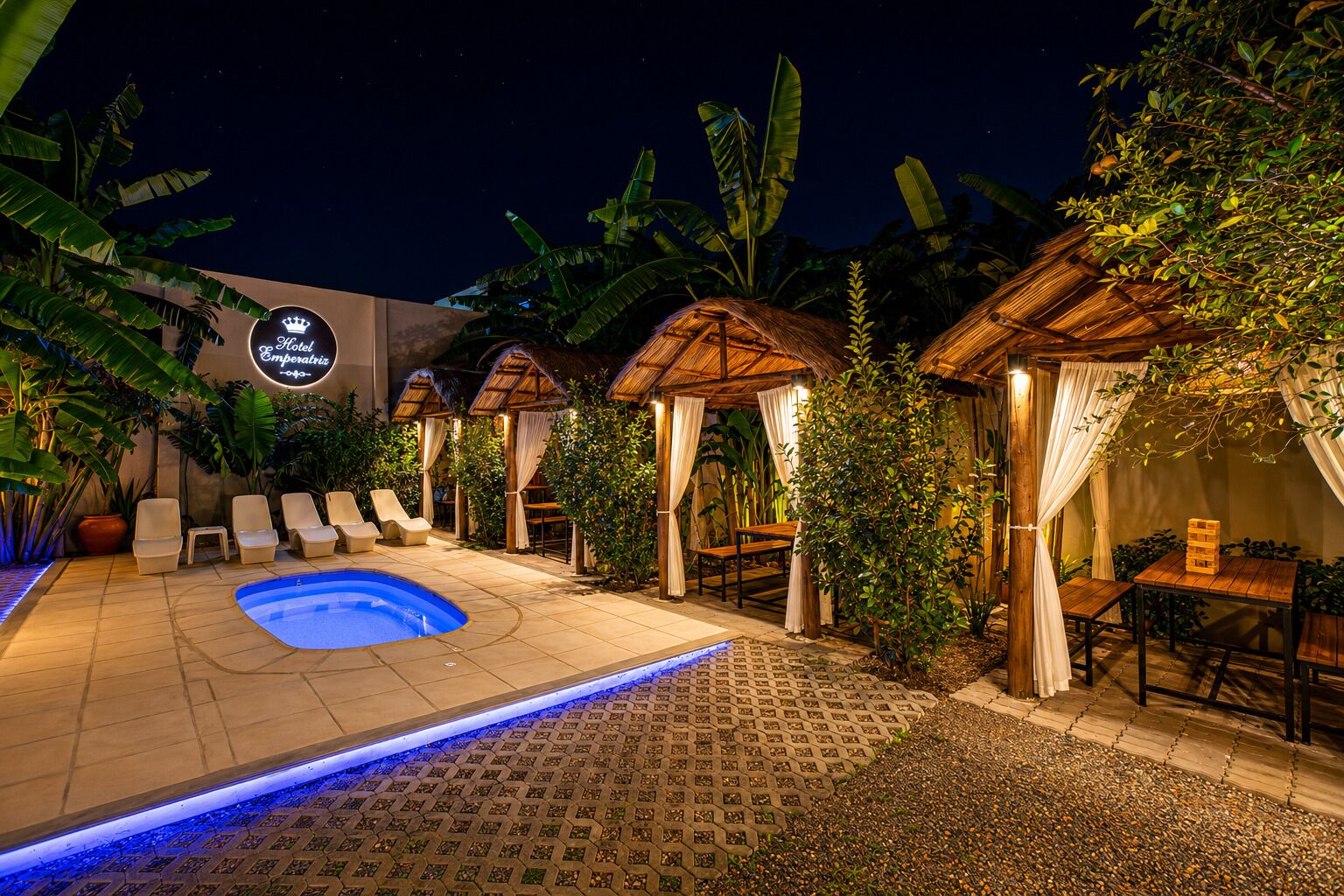 A hotel courtyard with a swimming pool, lounge chairs, and thatched-roof structures.