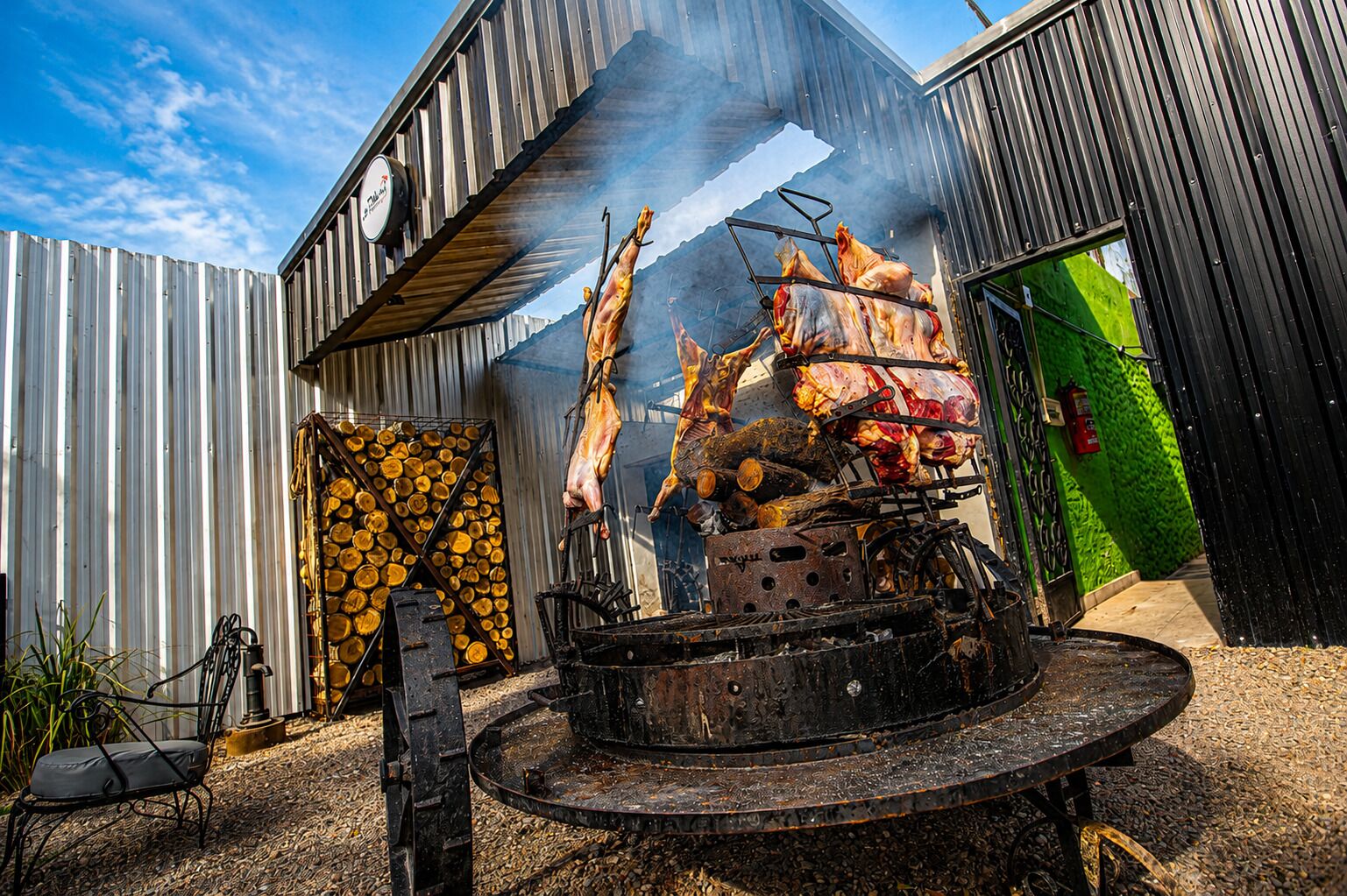 Een rustieke buitenkookopstelling met een metalen barbecue, een stapel hout en een groene wand.