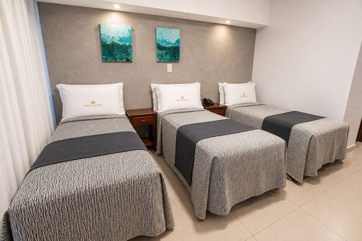 A hotel room with three beds, each with a grey patterned bedspread and white pillows, a wooden nightstand, and two abstract paintings on the wall.