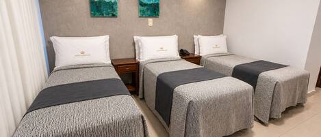 A hotel room with three beds, each with a grey patterned bedspread and white pillows, a wooden nightstand, and two abstract paintings on the wall.