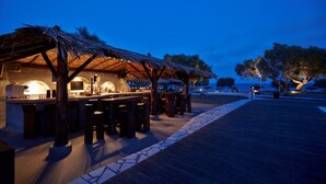 Area bar all'aperto con tetto di paglia, sgabelli di legno e pavimento in pietra.