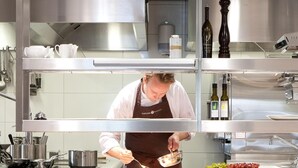Een kok in een professionele keuken, met roestvrijstalen apparatuur, een pan op het aanrecht en een fles wijn in de kast.
