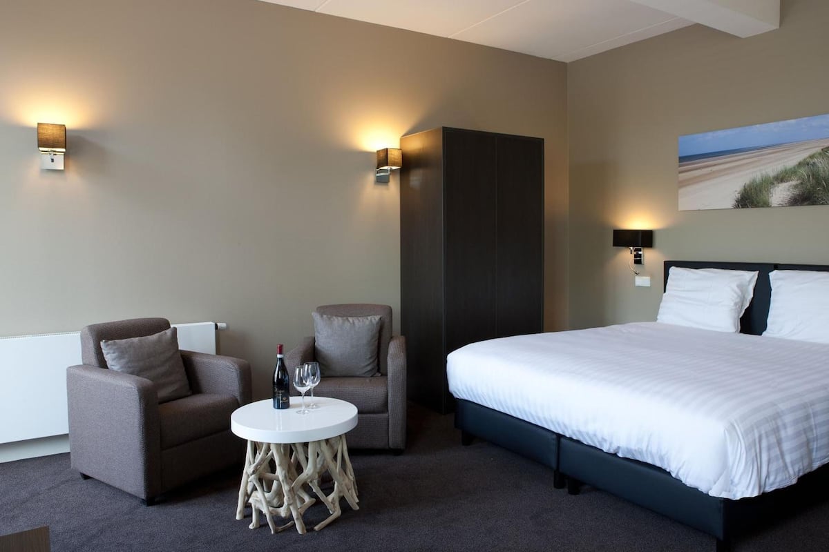 Une chambre d’hôtel avec un grand lit, deux fauteuils, une petite table ronde avec une bouteille et un verre, et deux lampes fixées au mur.