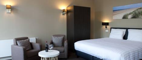 A hotel room with a large bed, two armchairs, a small round table with a bottle and glass, and two wall-mounted lights.