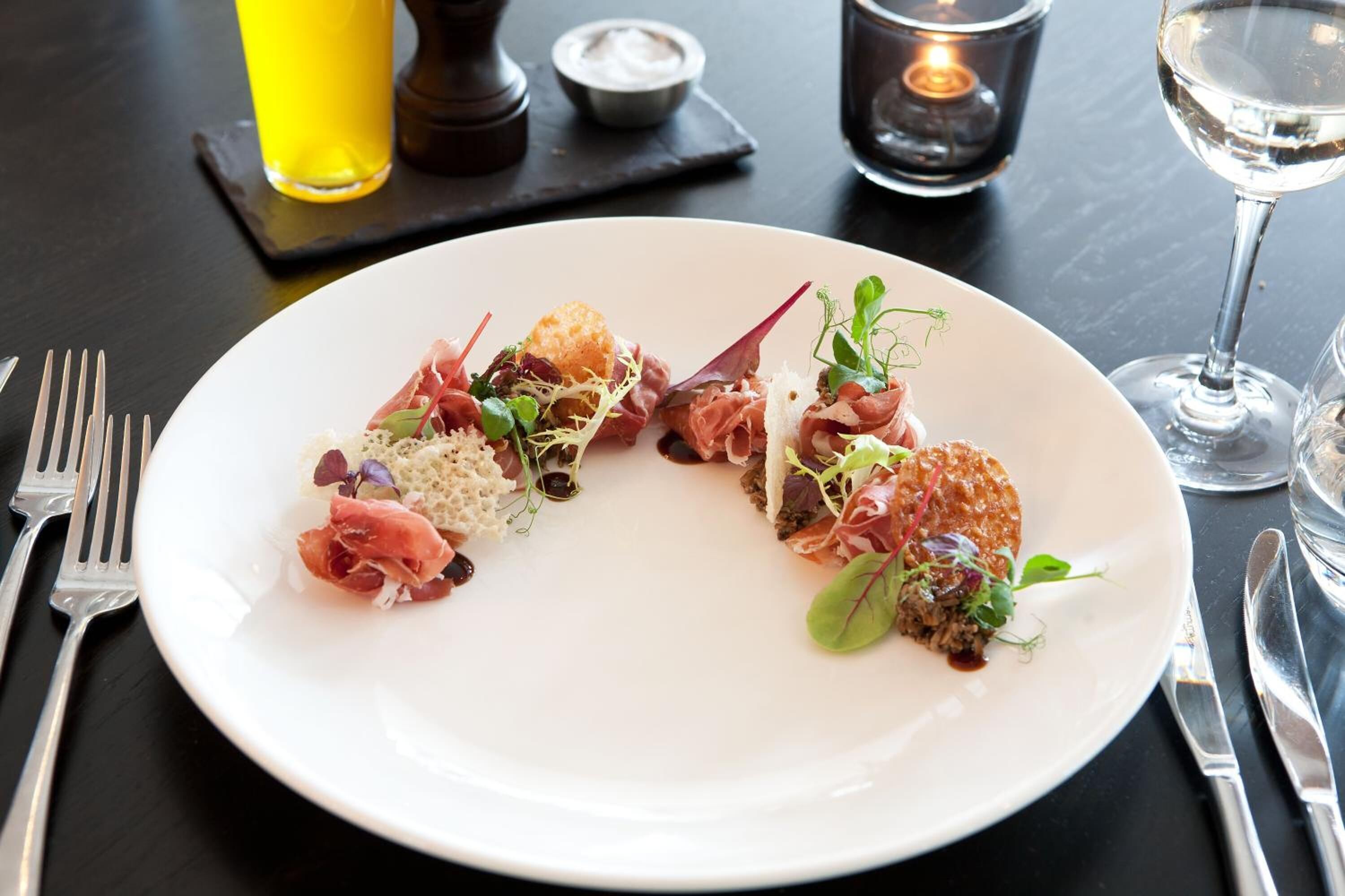 A white plate with a variety of food items, including prosciutto, greens, and a small dish of sauce, on a dark table with cutlery and a glass of water.