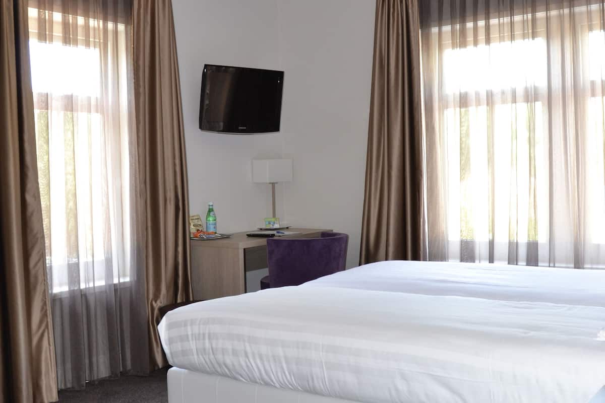 Une chambre d’hôtel avec un grand lit, un bureau avec une lampe, un téléviseur fixé au mur et des rideaux aux fenêtres.