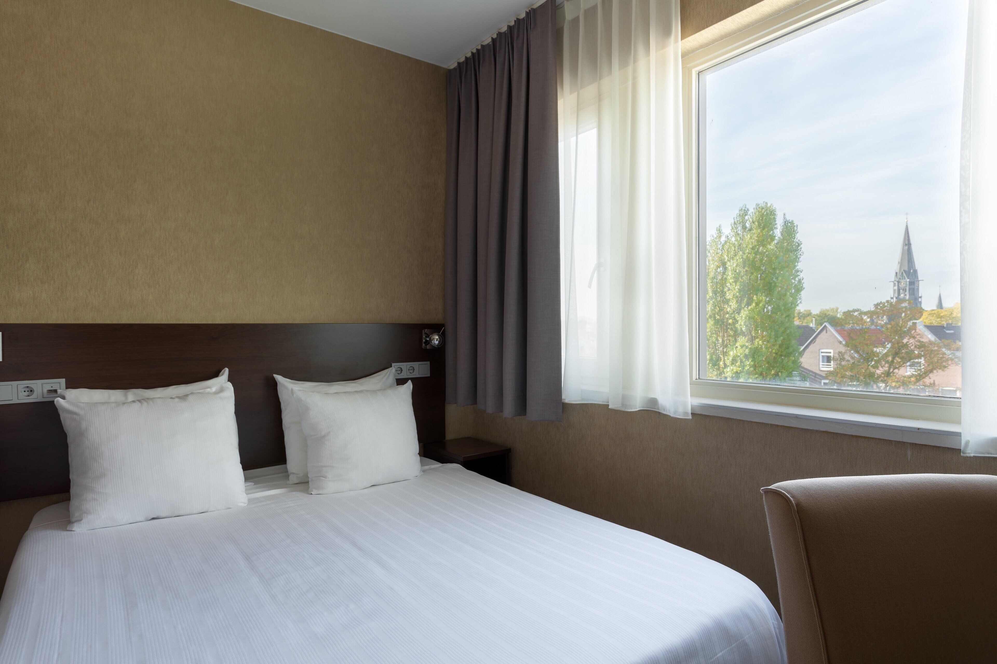 A hotel room with a large bed, two pillows, a nightstand, and a window with a view of a building and trees.