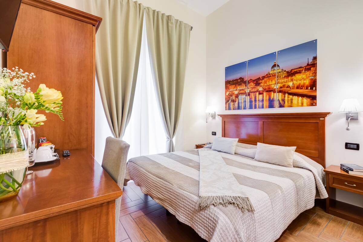 A hotel room with a bed, a desk with a vase of flowers, a painting of a cityscape, and a nightstand with a lamp.