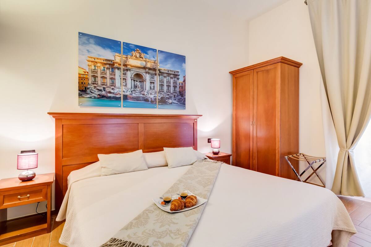A hotel room with a large bed, a wooden wardrobe, a nightstand with a lamp, and a wall-mounted painting of a building.