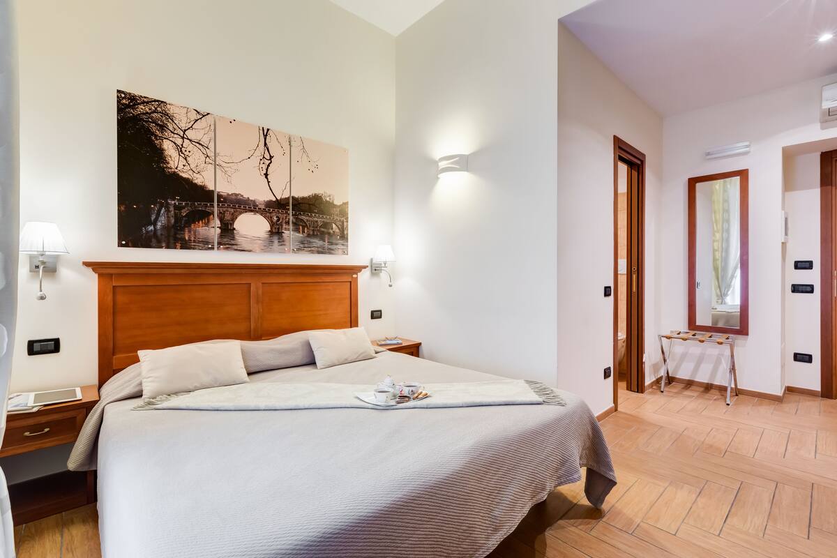 A neatly arranged hotel room with a large bed, a nightstand, a wall-mounted lamp, and a painting of a bridge.