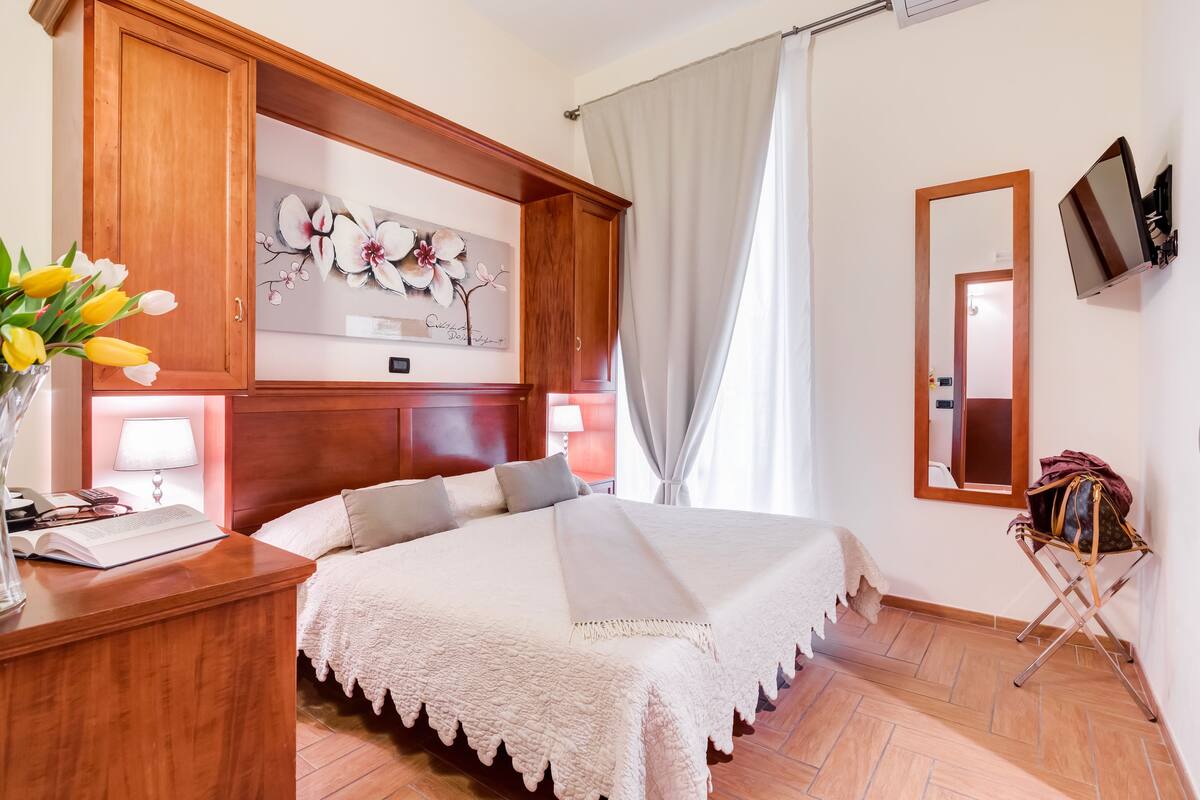 A hotel room with a large bed, wooden headboard, a desk with a lamp, a mirror, and a wall-mounted TV.