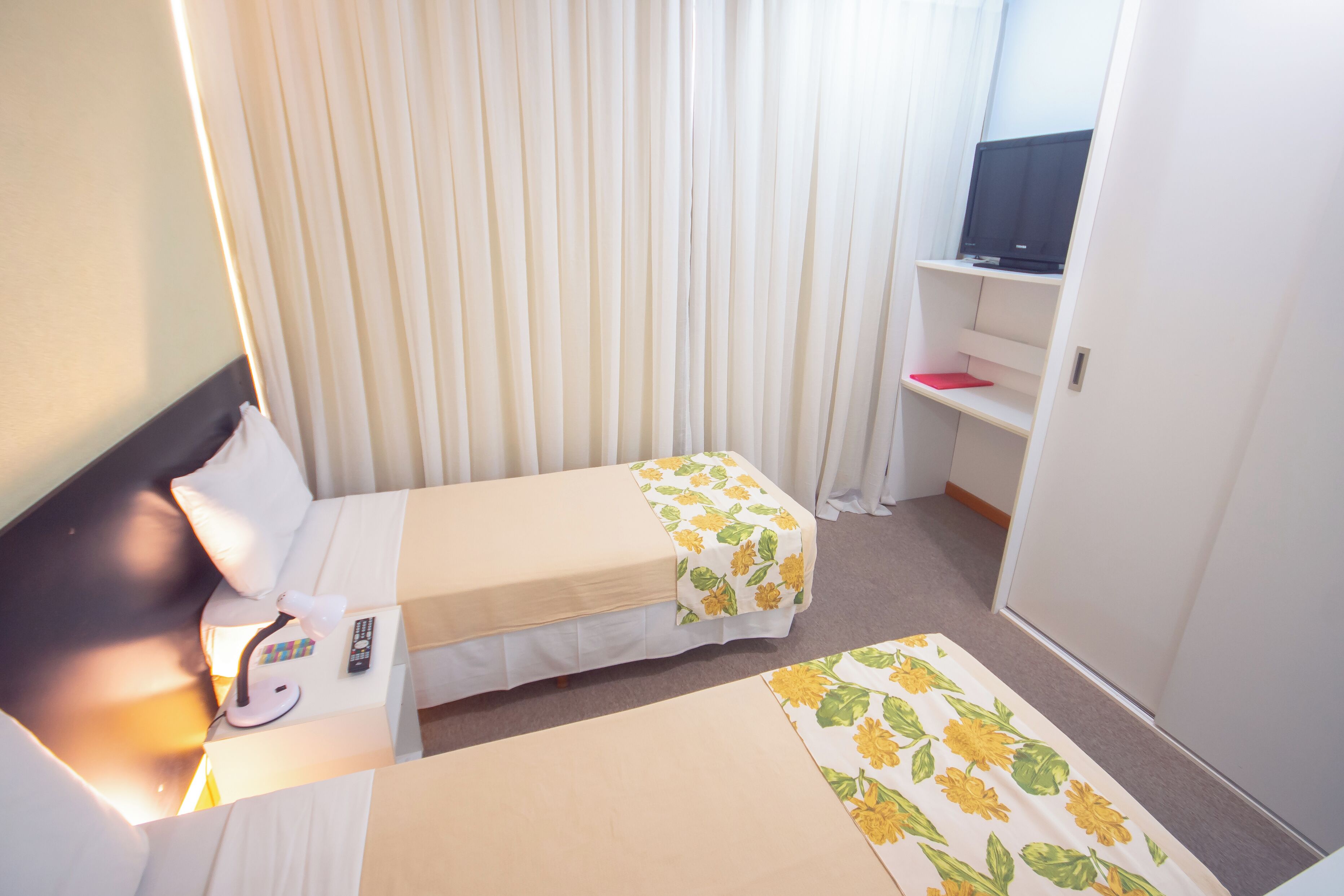 A hotel room with two single beds, a desk with a chair, a TV on a shelf, and a wardrobe.