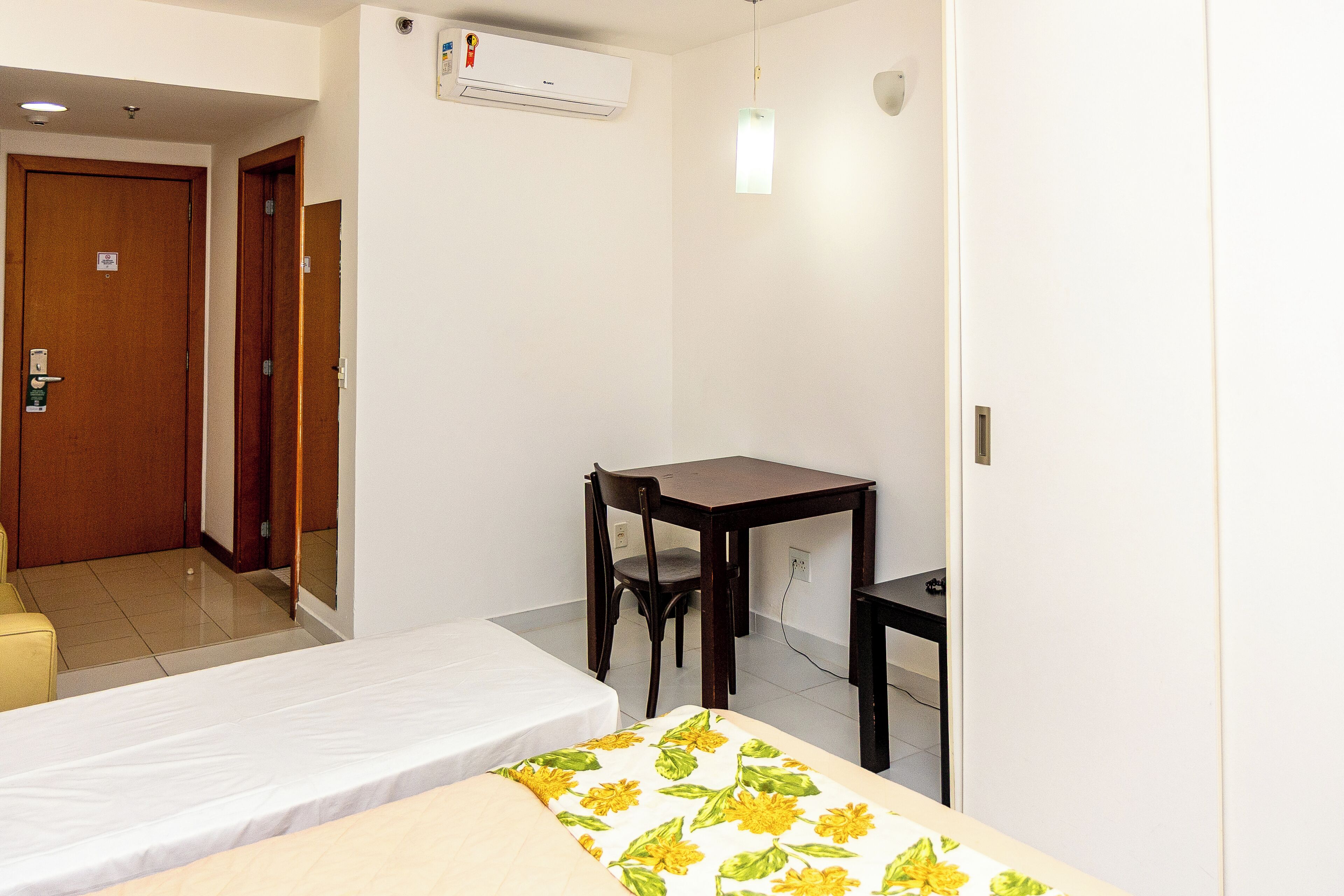 Une chambre d’hôtel avec un lit, un bureau et une chaise. Il y a un climatiseur fixé au mur et un luminaire au-dessus du bureau.
