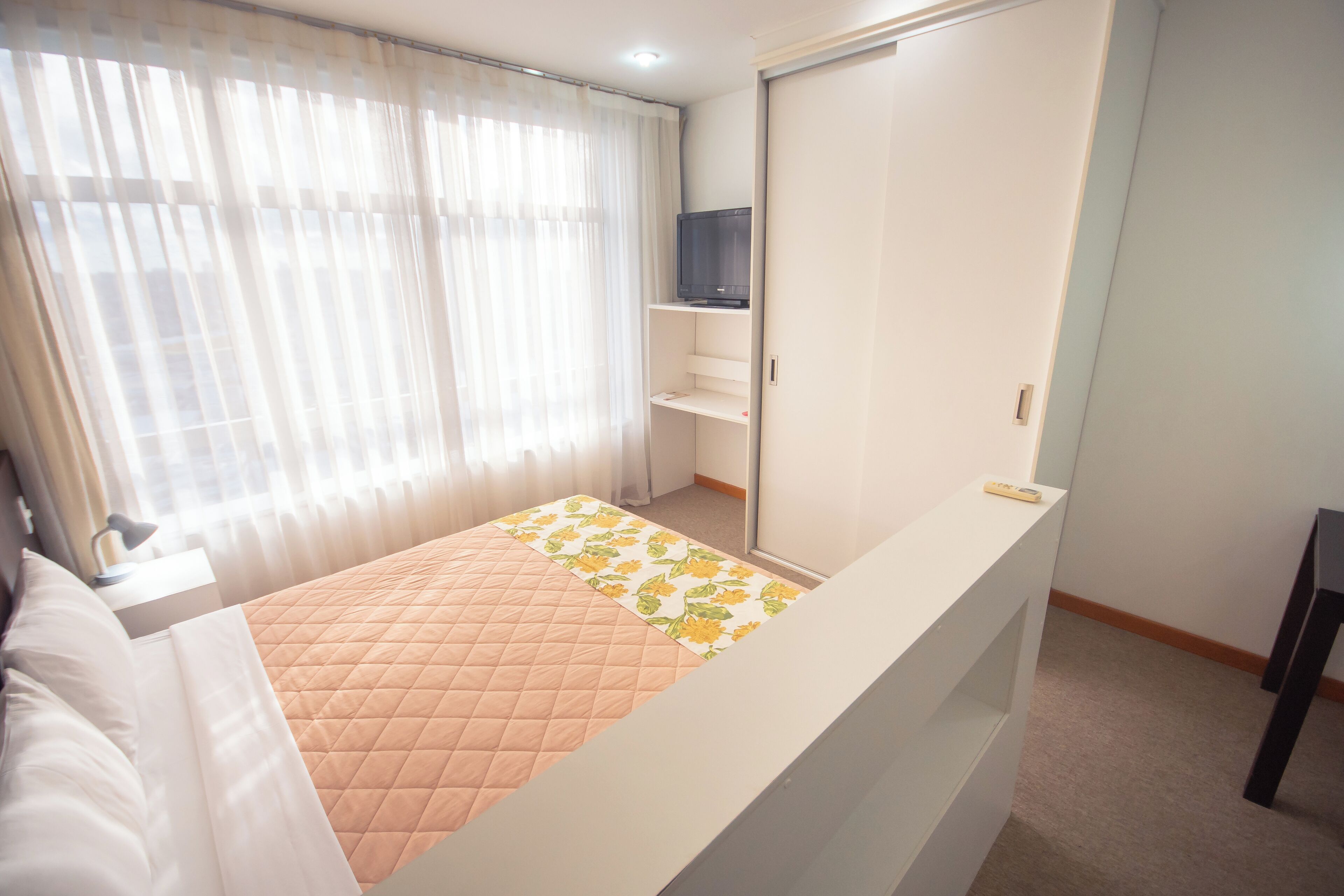 A bedroom with a bed, a TV on a shelf, a wardrobe, and a window with curtains.
