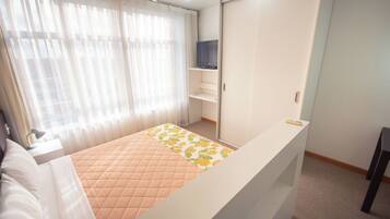 A bedroom with a bed, a TV on a shelf, a wardrobe, and a window with curtains.