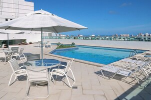 Un área de piscina en la azotea con sillones de descanso y sombrillas, con vistas despejadas del horizonte de la ciudad y un suelo de piscina con baldosas.