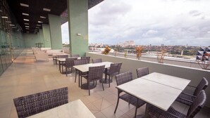 An outdoor seating area with tables and chairs, a glass railing, and a view of a cityscape.