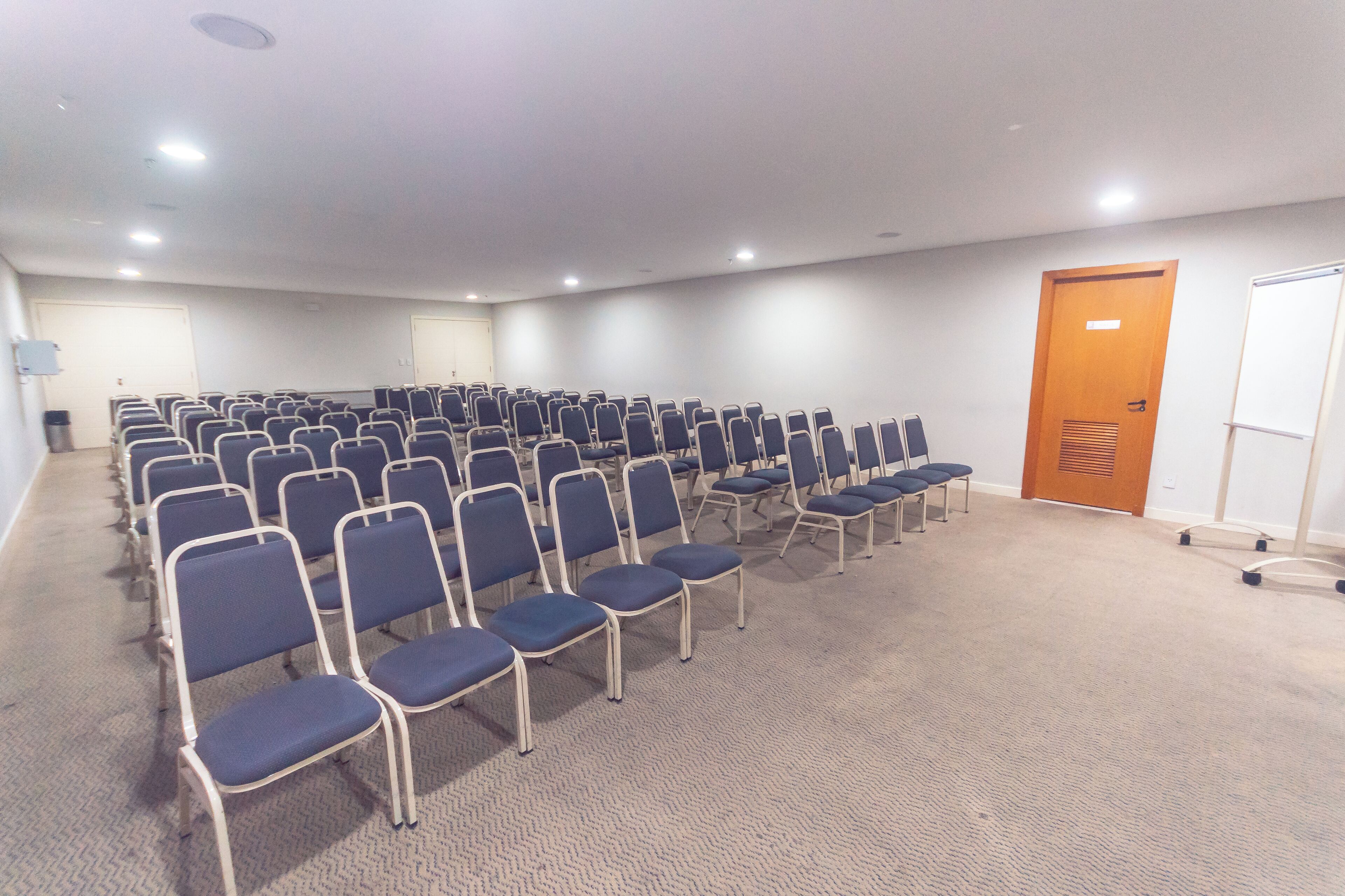 Une salle de conférence avec des rangées de chaises, un sol recouvert de moquette, des murs blancs et une porte en bois.