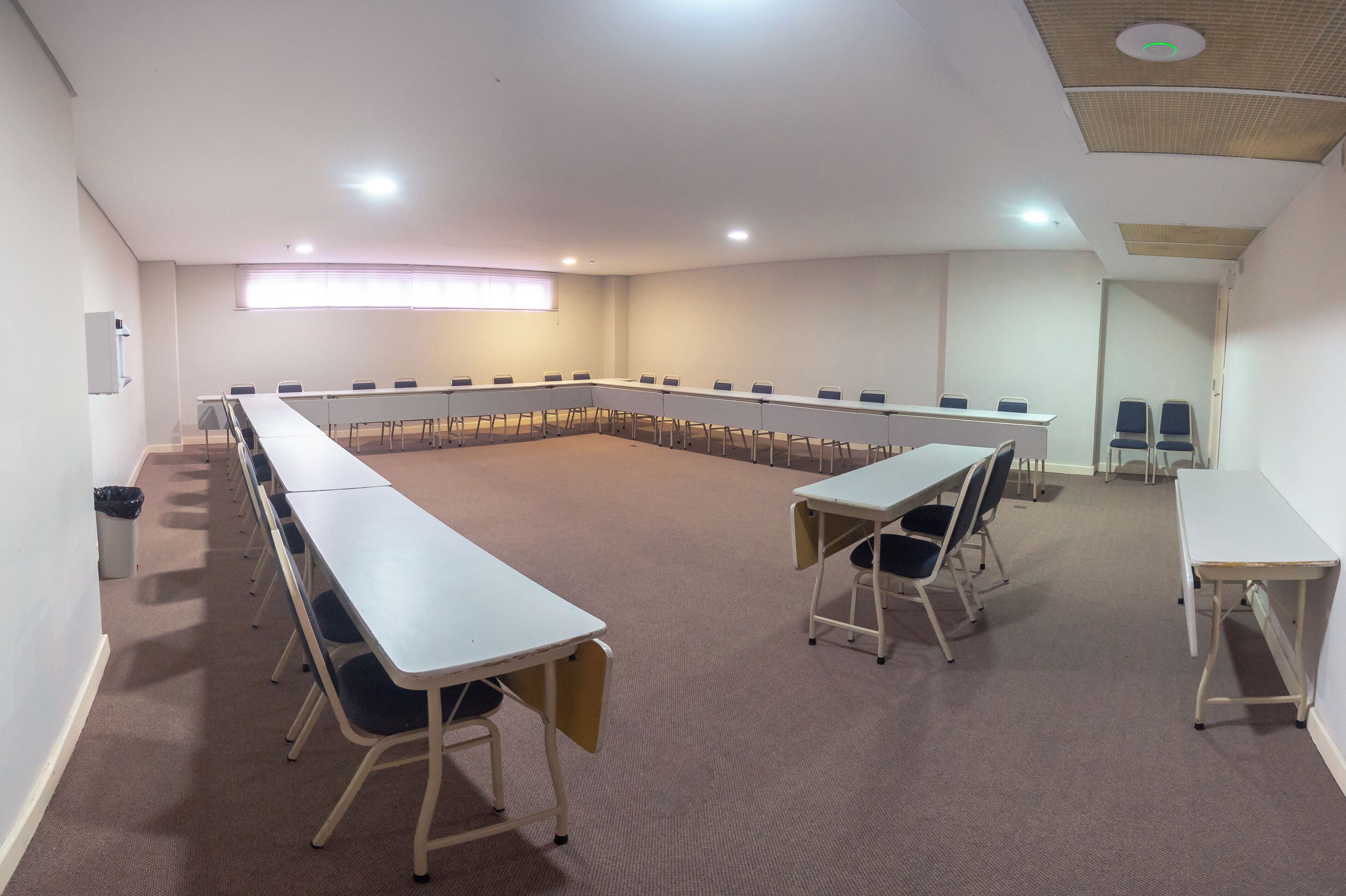Sala de conferência com uma mesa comprida e cadeiras, piso coberto de carpete e luminárias no teto.